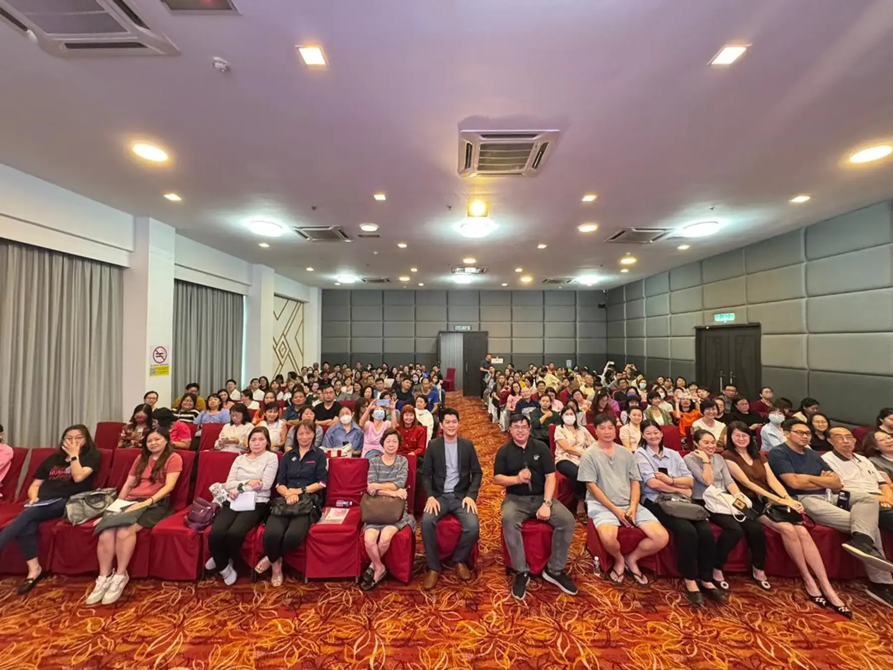 A large audience seated in a red-carpeted hall at a BBH seminar